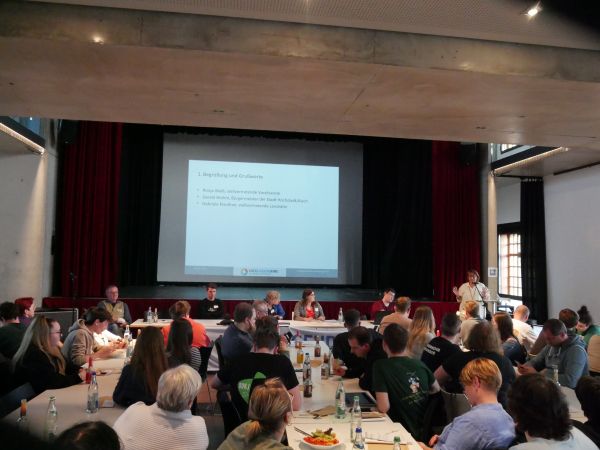 In einem großen Saal sitzen zahlreiche Menschen an mehreren Tischen, die mit Getränkeflaschen und Papieren bedeckt sind. Im Hintergrund befindet sich eine Bühne mit rotem Vorhang, auf der mehrere Personen an einem langen Tisch sitzen. Eine Person steht rechts am Rand der Bühne und spricht in ein Mikrofon. Im Zentrum der Bühne wird eine Präsentation auf eine Leinwand projiziert, deren Text teilweise lesbar ist. Die Atmosphäre wirkt konzentriert und geschäftig.