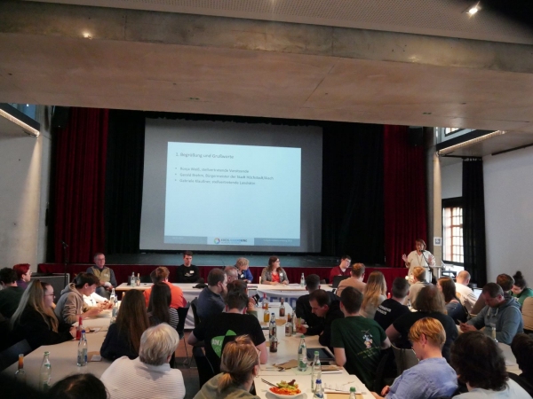 In einem großen Saal sitzen zahlreiche Menschen an mehreren Tischen, die mit Getränkeflaschen und Papieren bedeckt sind. Im Hintergrund befindet sich eine Bühne mit rotem Vorhang, auf der mehrere Personen an einem langen Tisch sitzen. Eine Person steht rechts am Rand der Bühne und spricht in ein Mikrofon. Im Zentrum der Bühne wird eine Präsentation auf eine Leinwand projiziert, deren Text teilweise lesbar ist. Die Atmosphäre wirkt konzentriert und geschäftig.