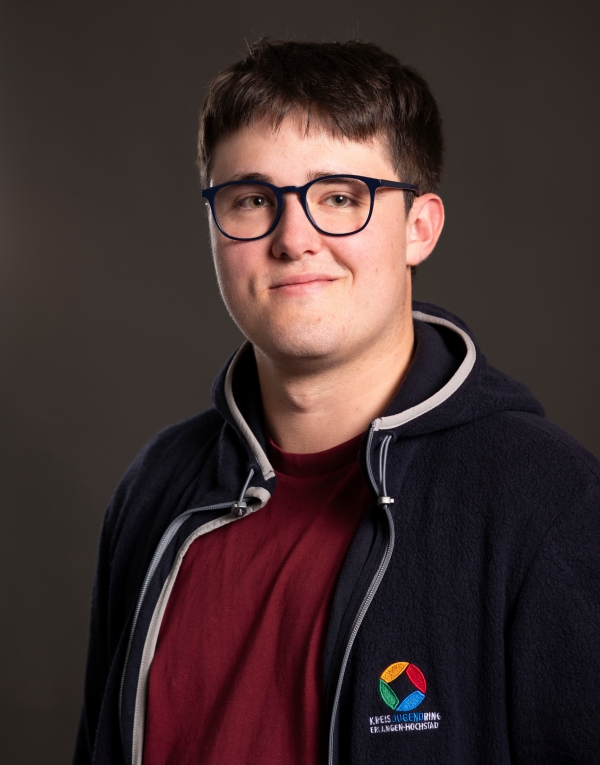 Junger Mann mit kurzem Haar trägt eine dunkelblaue Jacke mit farbigem Logo und ein rotes T-Shirt vor dunklem Hintergrund