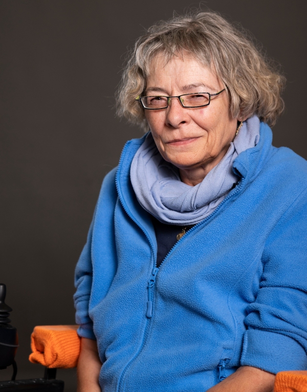 Ältere Person mit lockigem Haar trägt eine blaue Fleecejacke und einen hellblauen Schal vor dunklem Hintergrund