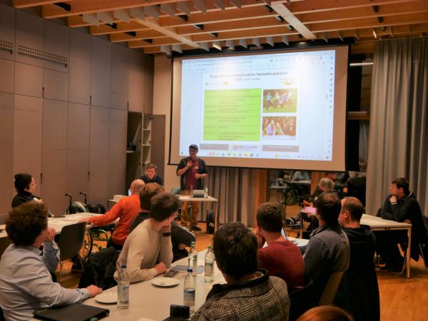 Mehrere Personen sitzen an Tischen in einem Konferenzraum. Im Vordergrund sind Menschen im Gespräch vertieft, einige machen sich Notizen oder schauen nach vorn. Im Hintergrund steht eine Person an einem Rednerpult und spricht in ein Mikrofon. Dahinter ist eine große Leinwand mit einer Präsentationsfolie zu sehen, die überwiegend grün gestaltet ist und Text sowie mehrere Fotos enthält. Die Decke besteht aus sichtbaren Holzbalken, an der rechten Seite hängt ein grauer Vorhang.