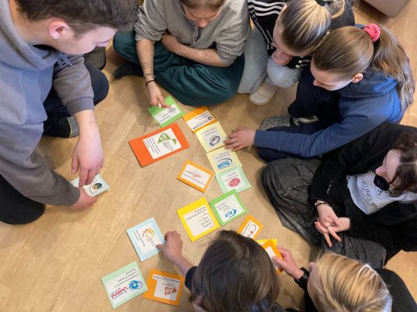 Mehrere Personen sitzen auf dem Boden in einem Kreis. Sie tragen legere Kleidung und beugen sich nach vorne, um bunte Karten und Umschläge zu betrachten, die auf dem hellen Holzfußboden verteilt sind. Die Karten zeigen verschiedene Symbole, Logos und Schriftzüge, die teilweise gut lesbar sind. Die Szene vermittelt einen konzentrierten, gemeinschaftlichen Moment.