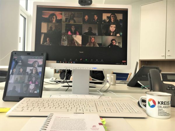 Ein Schreibtisch mit einem Computerbildschirm, auf dem eine Videokonferenz mit mehreren Teilnehmenden zu sehen ist. Links steht ein Tablet, das ebenfalls eine Videokonferenz zeigt. Im Vordergrund liegt ein aufgeschlagenes Heft mit Text und Grafiken. Rechts steht eine weiße Tasse mit mehrfarbigem Logo und der Aufschrift 'KREISJUGENDRING ERLANGEN-HÖCHSTADT'. Im Hintergrund sind weiße Büromöbel und ein Fenster erkennbar.