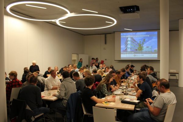 In einem modernen Konferenzraum sitzen zahlreiche Menschen an langen, hellen Tischen. Die Personen sind in angeregten Gesprächen vertieft oder schauen auf ihre Unterlagen. An der Wand hängt eine große Leinwand mit einer Präsentation, die ein Gebäude und einen Text zeigt. Die Deckenbeleuchtung besteht aus zwei kreisförmigen Lichtinstallationen. Die Atmosphäre wirkt geschäftig und konzentriert.