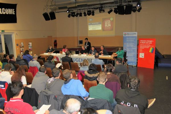 In einem Saal sitzen zahlreiche Personen auf Stühlen und blicken nach vorn zu einem Podium, an dem mehrere Menschen hinter Tischen sitzen. Auf dem Podium stehen Mikrofone und Namensschilder. Im Hintergrund hängen mehrere Banner und Roll-ups mit Text und Logos, darunter ein großes Banner mit der Aufschrift 'WELCOME'. Die Wände sind hell gestrichen, und an der Decke sind Scheinwerfer und Lautsprecher angebracht. Die Szene wirkt wie eine Podiumsdiskussion oder Informationsveranstaltung.