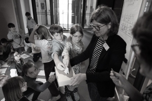 Gruppe von Kindern, die in einem Raum an einer Wahl oder Abstimmung teilnehmen, eine Frau hält eine Box, in die die Kinder Zettel einwerfen