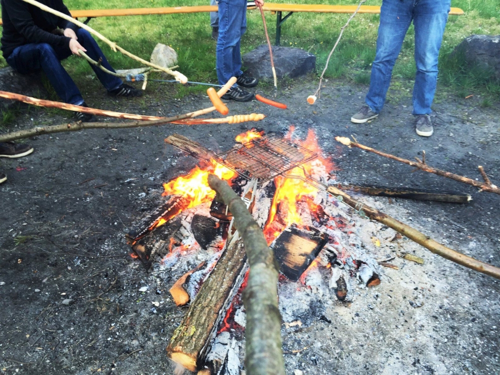 Mehrere Personen stehen oder sitzen um ein Lagerfeuer auf einer offenen Fläche. Über dem Feuer liegen Holzscheite und ein Metallrost. Verschiedene lange Stöcke mit Würstchen und Marshmallows werden von außen ins Feuer gehalten. Im Hintergrund sind Gras, Steine und eine Bank zu sehen. Die Szene vermittelt eine entspannte Atmosphäre beim gemeinsamen Grillen im Freien.
