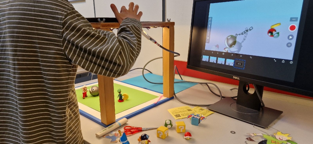 Ein Kind mit gestreiftem Langarmshirt steht an einem Tisch und arbeitet an einem Trickfilmprojekt. Auf dem Tisch stehen eine kleine Holzrahmen-Konstruktion mit Kamera, eine grüne Unterlage, mehrere Spielfiguren und Requisiten aus Kunststoff sowie Bastelmaterial wie Schere und Papier. Ein Monitor zeigt eine Animationsszene mit Spielfiguren und Werkzeugleiste einer Animationssoftware.