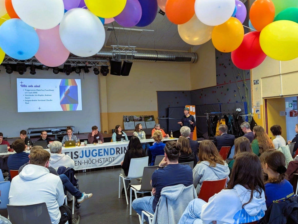 Bunte Luftballons hängen von der Decke eines großen Raumes. Im Vordergrund sitzen zahlreiche Menschen auf Stühlen und blicken zu einer Gruppe, die an einem langen Tisch auf einer Bühne sitzt. Hinter dem Tisch hängt ein Banner mit dem Schriftzug 'JUGENDRIN'. Links neben der Bühne ist eine Leinwand mit einer Präsentation zu sehen, auf der ein Text und ein farbiges Diagramm abgebildet sind. Die Wände sind hell gestrichen, und im Hintergrund sind Klettergriffe an einer Wand befestigt.
