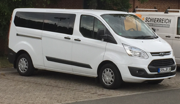 Weißer Kleinbus mit abgedunkelten Seitenscheiben und schwarzen Zierleisten steht auf einem gepflasterten Parkplatz. Die Frontpartie ist abgerundet, die Scheinwerfer sind schmal und seitlich angeordnet. Das Fahrzeug trägt ein Ford-Logo auf dem Kühlergrill. Im Hintergrund ist ein weiteres Fahrzeug mit Werbeaufschrift zu sehen.