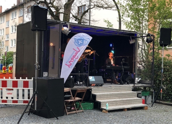 Eine mobile Bühne steht auf einem gepflasterten Platz. Die Bühne besteht aus einem schwarzen Anhänger mit ausklappbarer Front und Überdachung. Auf der Bühne sitzt eine Person an einem Keyboard und singt in ein Mikrofon, daneben steht eine Gitarre. Vor der Bühne führen silberne Stufen mit Riffelblech-Oberfläche hinauf. Links neben der Bühne ist eine weiße Fahne mit farbigem Logo und Schriftzug befestigt. Große Lautsprecher stehen rechts und links am Bühnenrand. Im Hintergrund sind Bäume und mehrstöckige Wohnhäuser zu sehen.