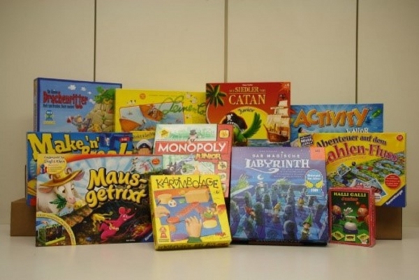 Verschiedene bunte Brettspiele und Kartenspiele stehen nebeneinander auf einer Holzfläche. Die Spieleverpackungen zeigen unterschiedliche Motive, darunter Illustrationen von Labyrinthen, Inseln, Tieren und Zahlen. Einige Schachteln sind größer und stehen hinten, kleinere Spiele sind vorne angeordnet. Die Titel und Logos der Spiele sind deutlich sichtbar, darunter 'Monopoly', 'Das magische Labyrinth', 'Make'n Break', 'Mausgetrix', 'Activity', 'Abenteuer auf dem Zahlenfluss', 'Halli Galli', 'Karambolage' und 'Die Siedler von Catan'.