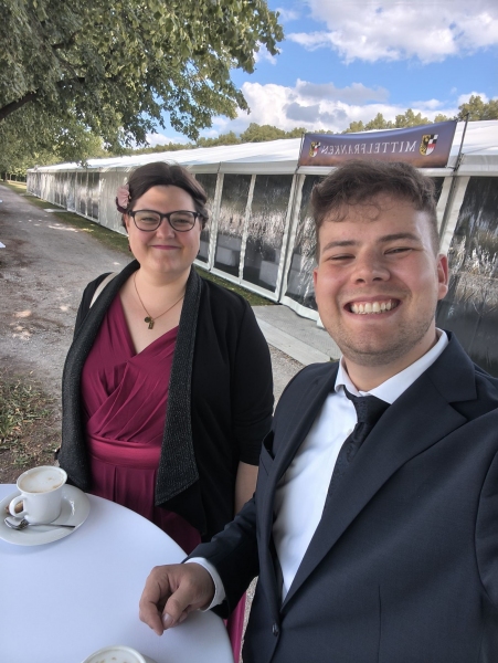 Ein Mann und eine Frau in festlicher Kleidung stehen an einem runden kleinen Tisch vor einem großen Festzelt unter einem Baum. Sie lächeln in die Kamera.